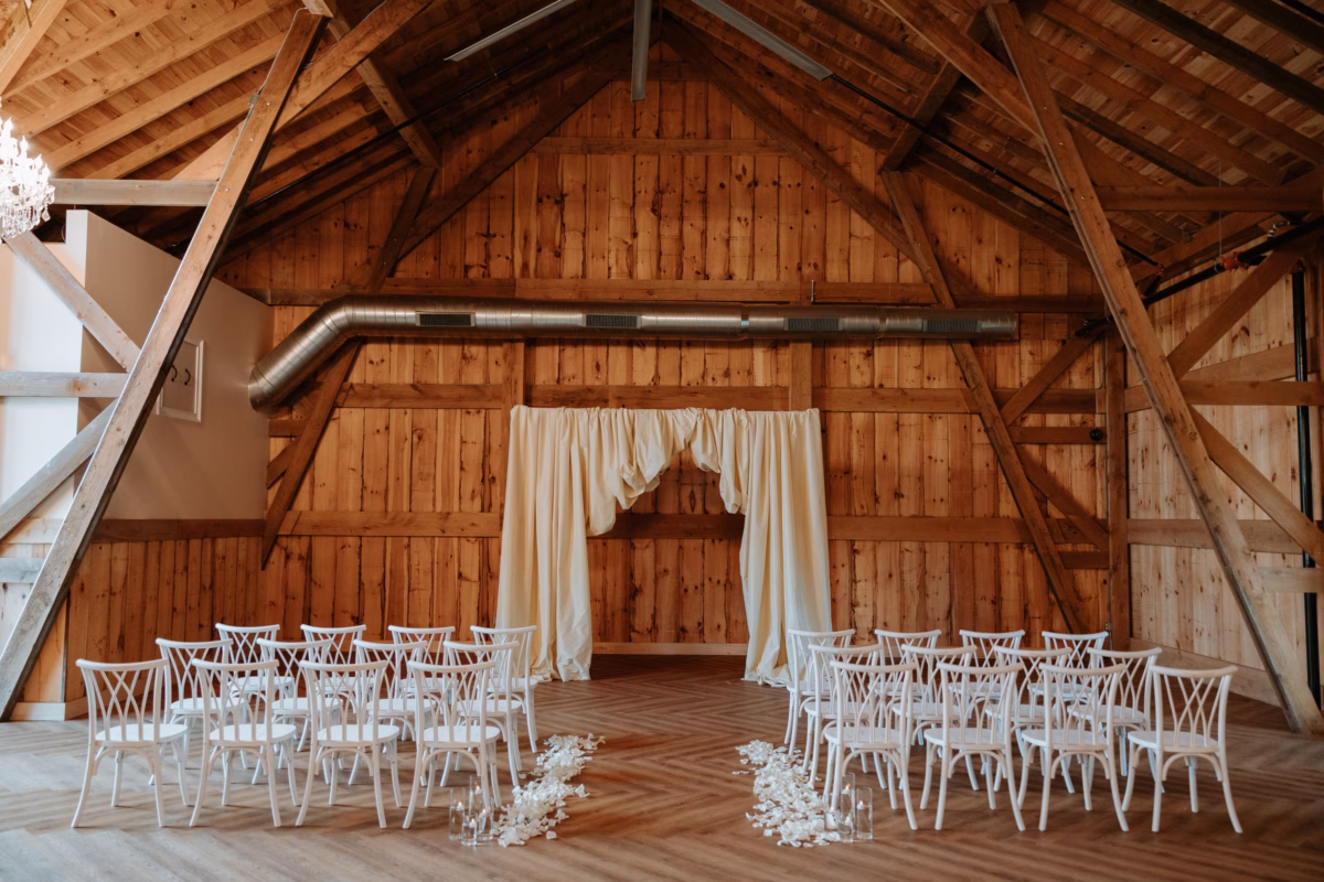 Rustic barn wedding ceremony space at The Grand in York SC near Charlotte NC featuring white crossback chairs arranged in rows with a floral petal aisle and draped cream fabric altar backdrop beneath exposed wooden beam cathedral ceiling