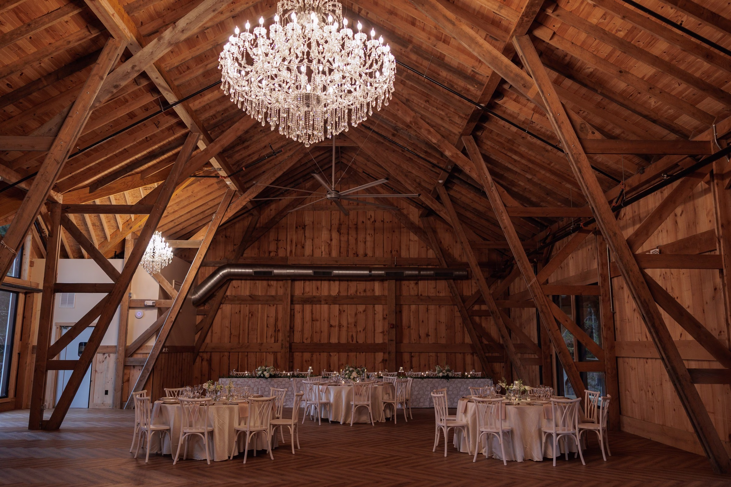 Rustic barn wedding reception hall at The Grand in York SC near Charlotte NC featuring soaring wood beam cathedral ceiling elegant crystal chandelier and white linen round tables set for dinner