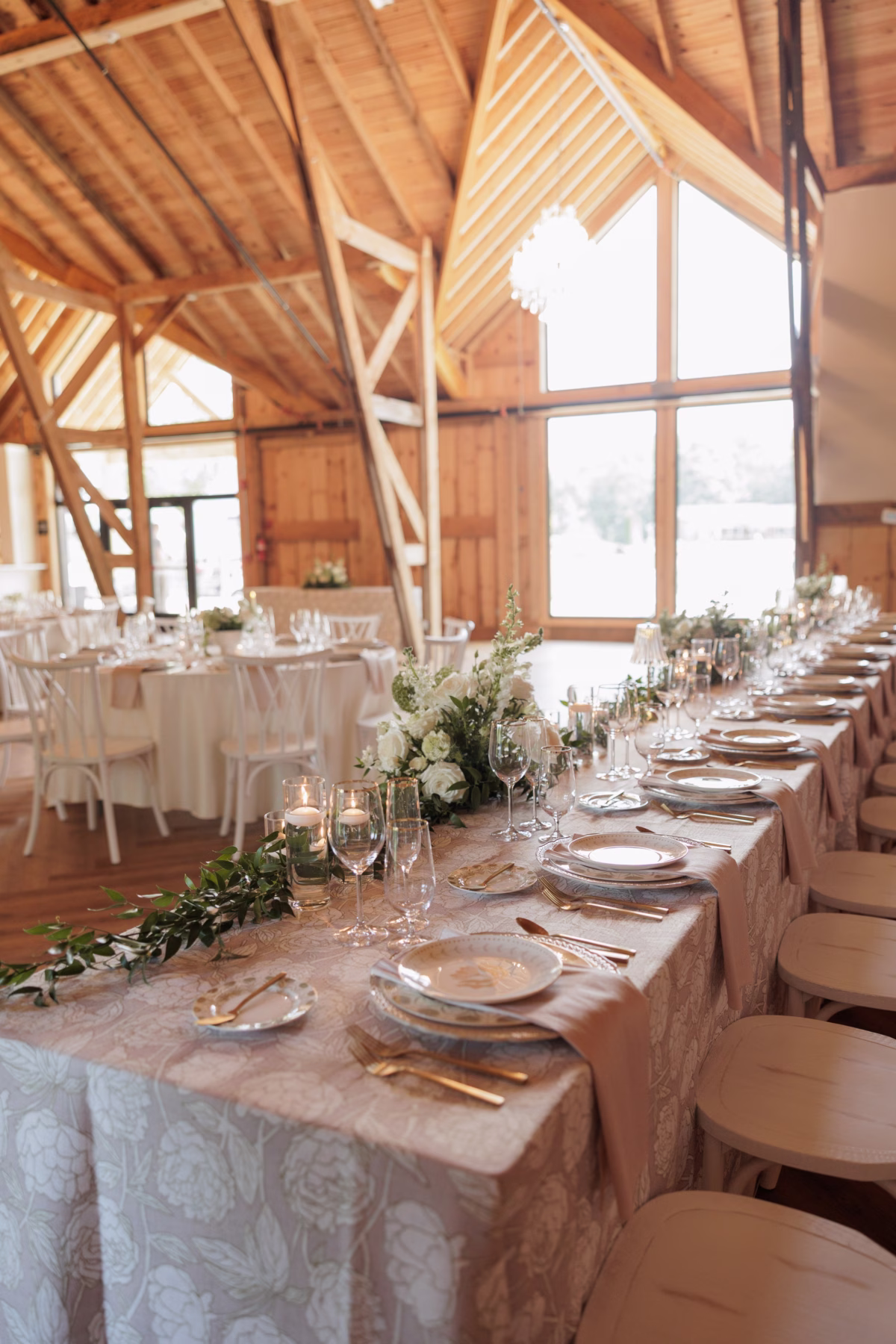 Elegant barn wedding reception at The Grand in York SC near Charlotte NC featuring a long head table with gold flatware, white floral greenery garland centerpieces, floating candles, and patterned blush linen tablecloth