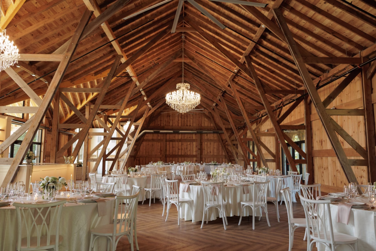 Rustic barn wedding reception at The Grand in York SC near Charlotte NC featuring exposed wooden beam cathedral ceiling crystal chandeliers white cross-back chairs and elegantly set round tables with floral centerpieces