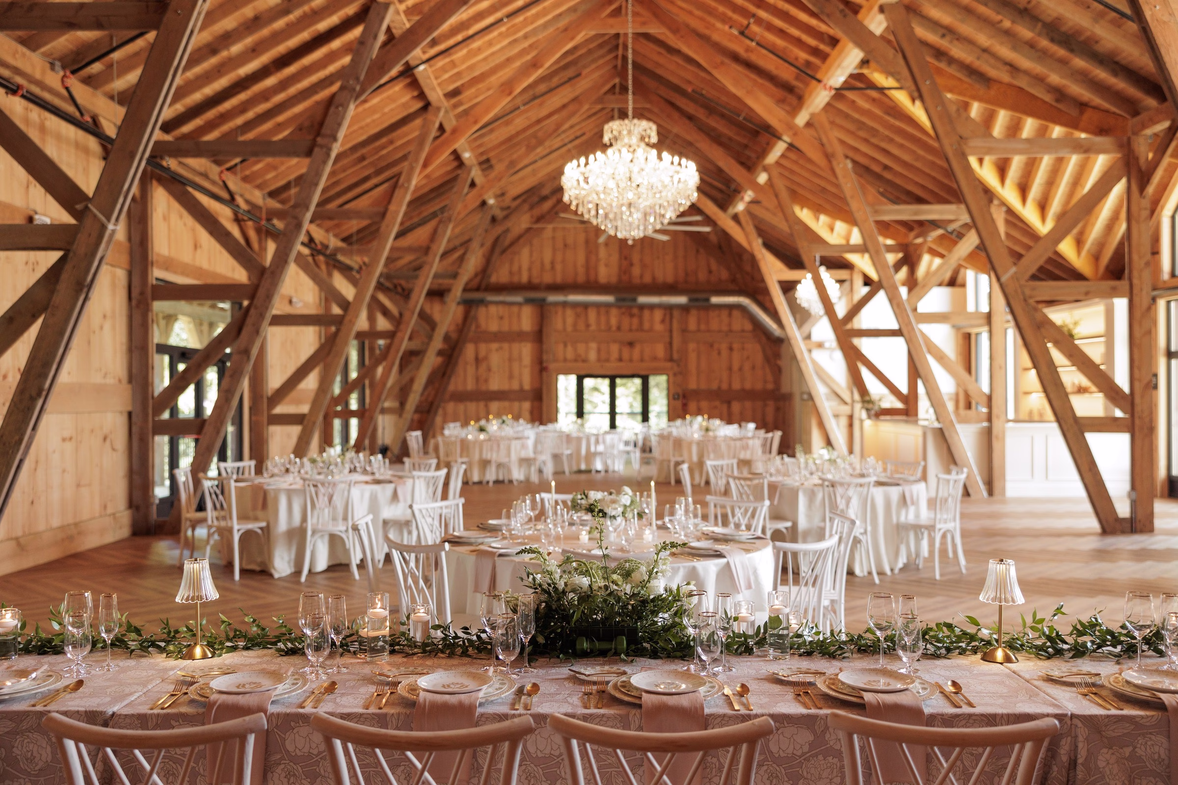 Elegantly decorated barn wedding reception at The Grand in York SC near Charlotte NC featuring a crystal chandelier exposed wood beam ceiling lush greenery garland runner gold flatware and white chiavari chairs at round and long tables