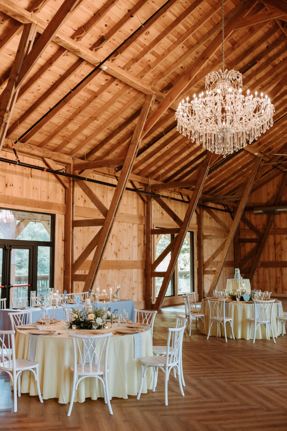 Elegant barn wedding reception at The Grand in York SC near Charlotte NC featuring a crystal chandelier soaring wood beam ceilings cream linen round tables and white crossback chairs