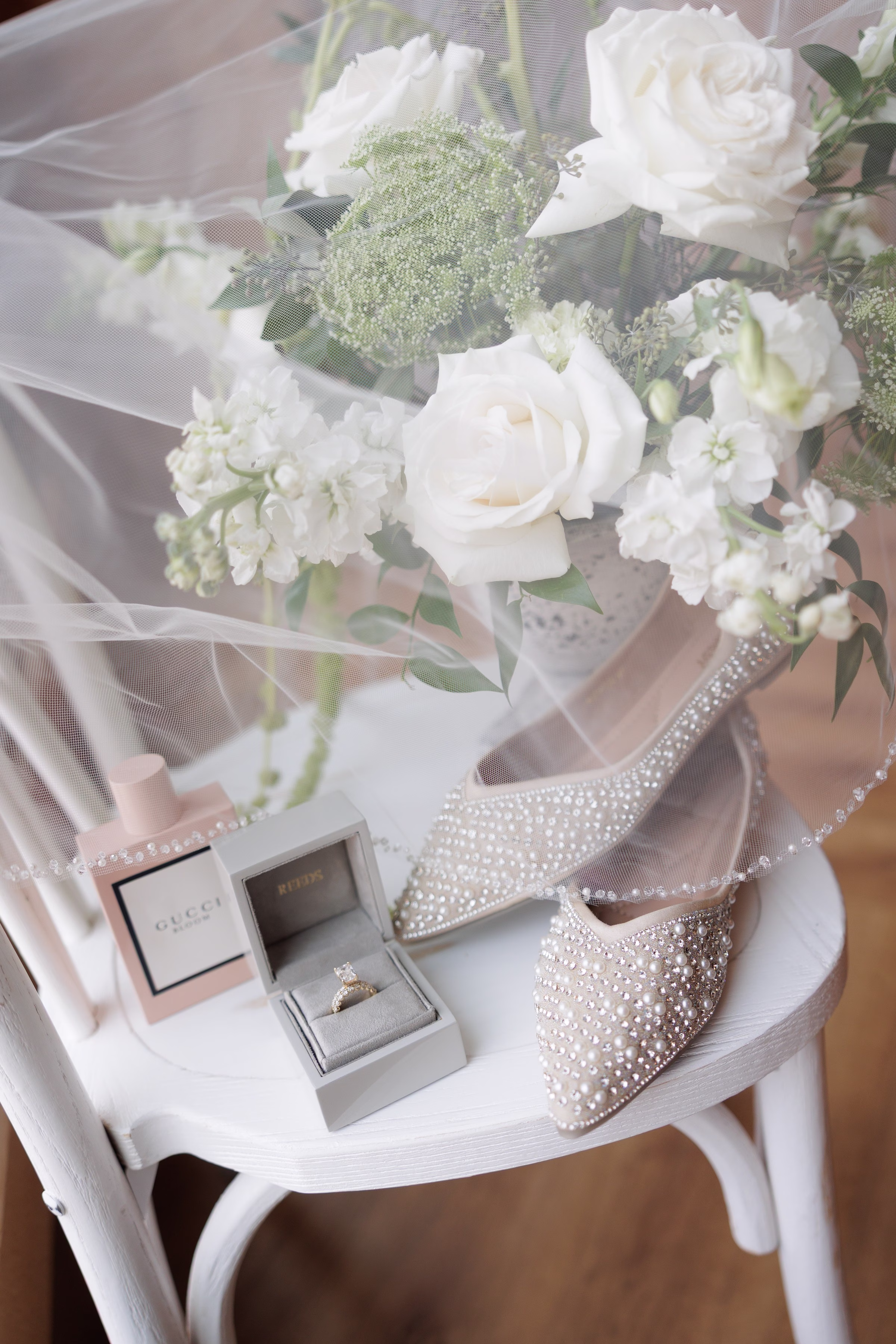 Bridal details flatlay featuring pearl-encrusted heels, diamond engagement ring in a Reeds Jewelers box, Gucci perfume, and a white rose bouquet on a white chair at The Grand wedding venue in York SC near Charlotte NC