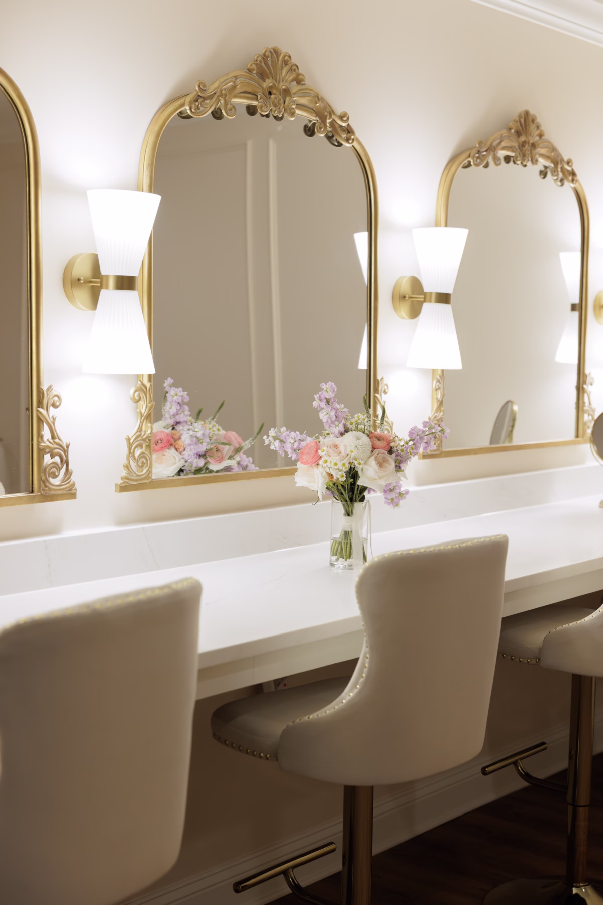 Elegant bridal suite vanity with ornate gold arched mirrors, gold sconce lighting, cream upholstered chairs, and a fresh floral arrangement at The Grand wedding venue in York SC near Charlotte NC