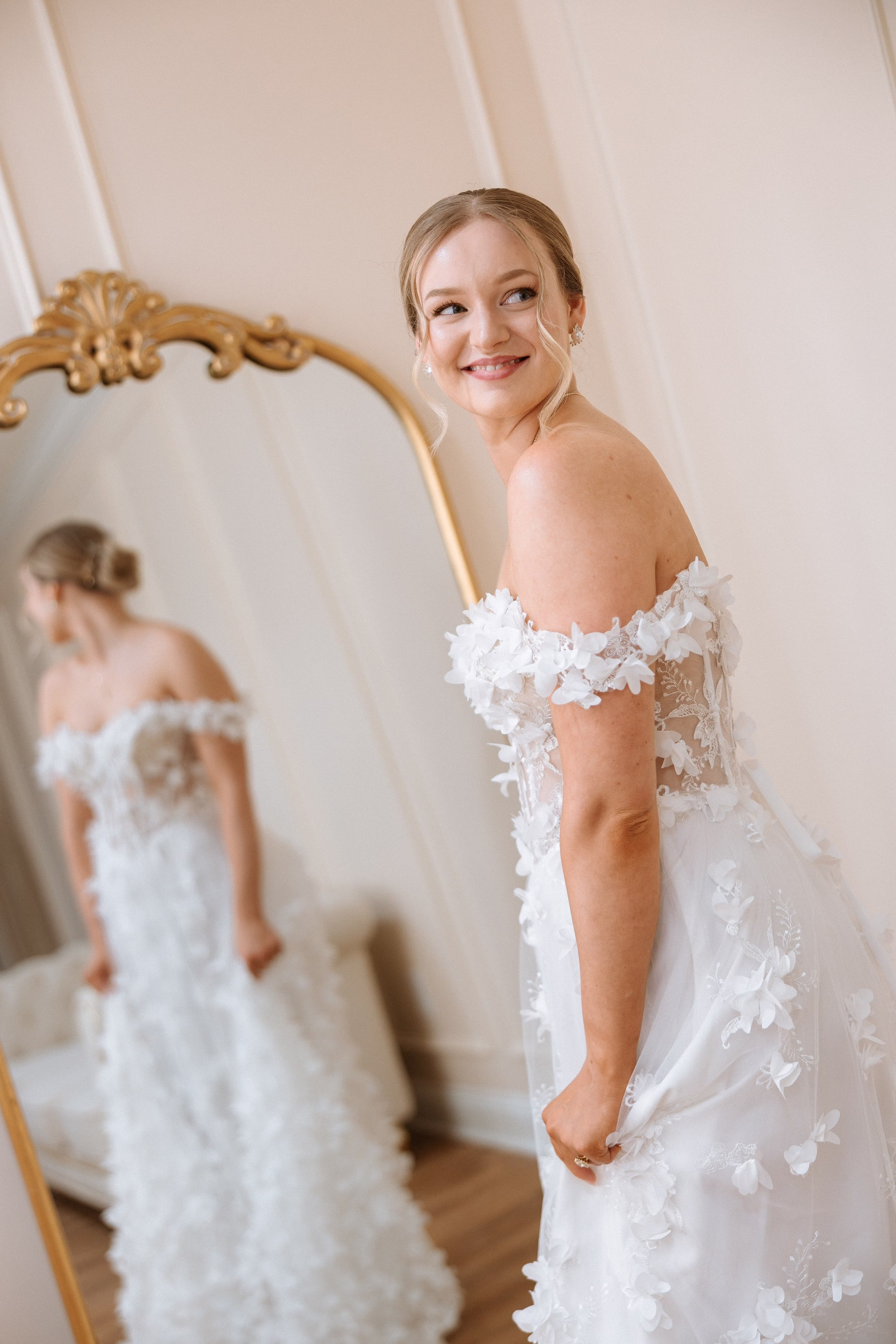 Smiling bride in an off-shoulder 3D floral applique wedding gown admiring her reflection in a gold ornate mirror at The Grand wedding venue in York SC near Charlotte NC