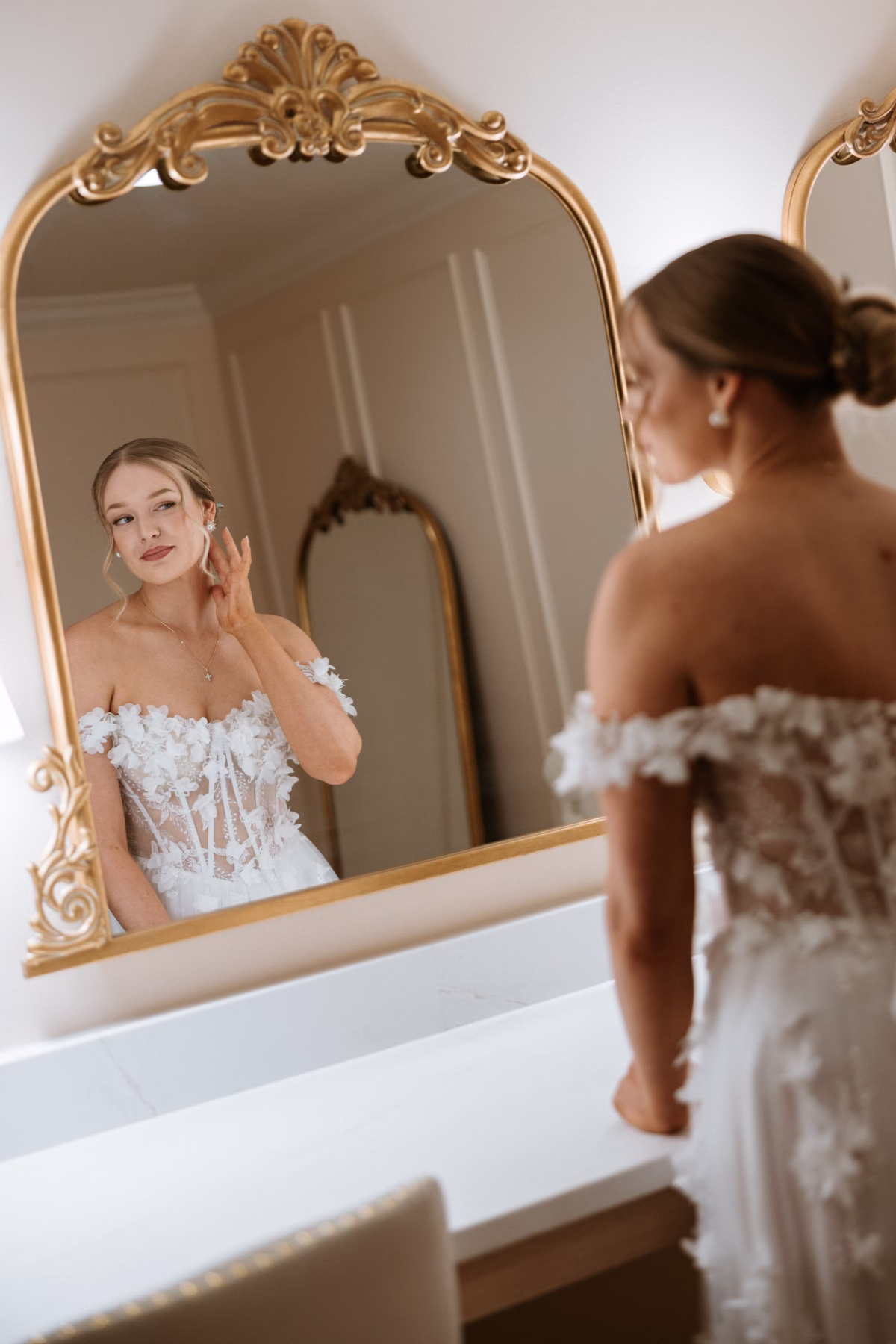 Bride in an off-shoulder floral lace wedding gown adjusting her earring while looking into an ornate gold mirror during bridal preparations at The Grand wedding venue in York SC near Charlotte NC