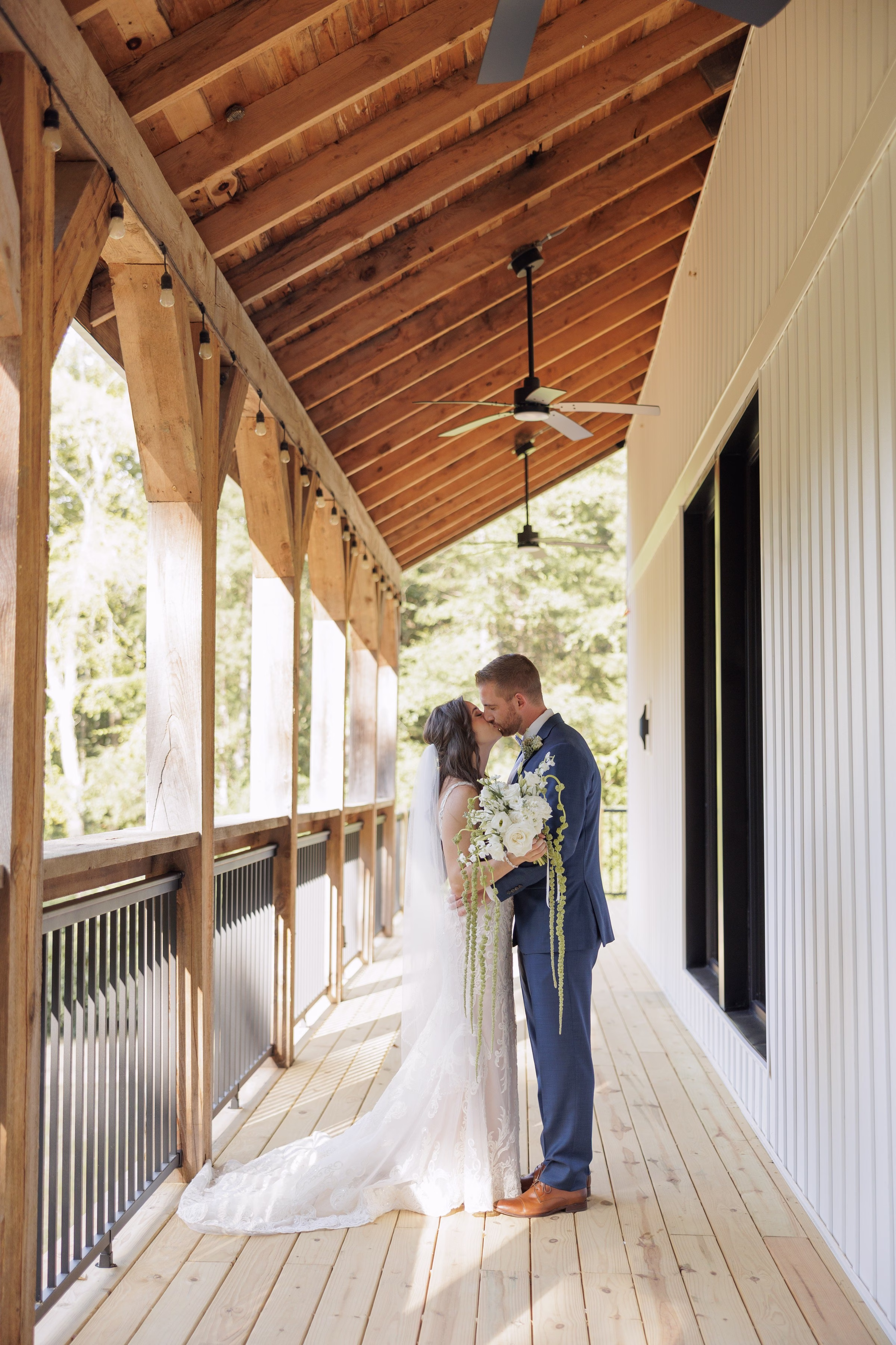 Bride and groom sharing a first kiss on the covered wooden porch at The Grand wedding venue in York SC near Charlotte NC, bride holding a cascading white floral bouquet with greenery while wearing a lace gown with cathedral train and groom in a navy suit