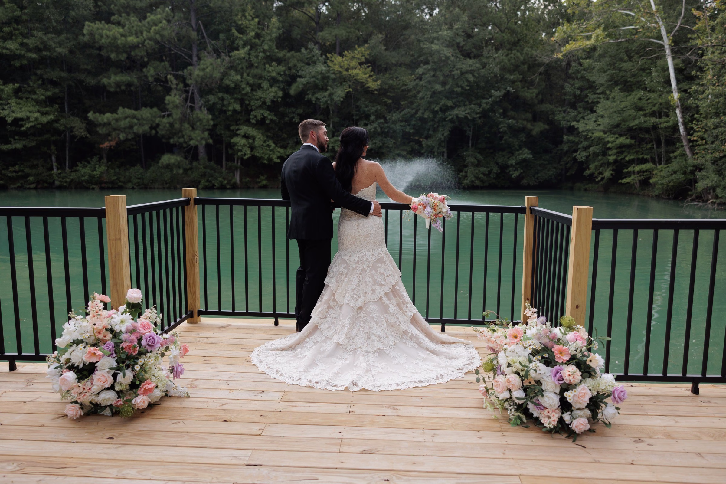 Bride in a lace mermaid gown and groom in a black tuxedo stand on a wooden deck overlooking a green lake surrounded by trees at The Grand wedding venue in York SC near Charlotte NC