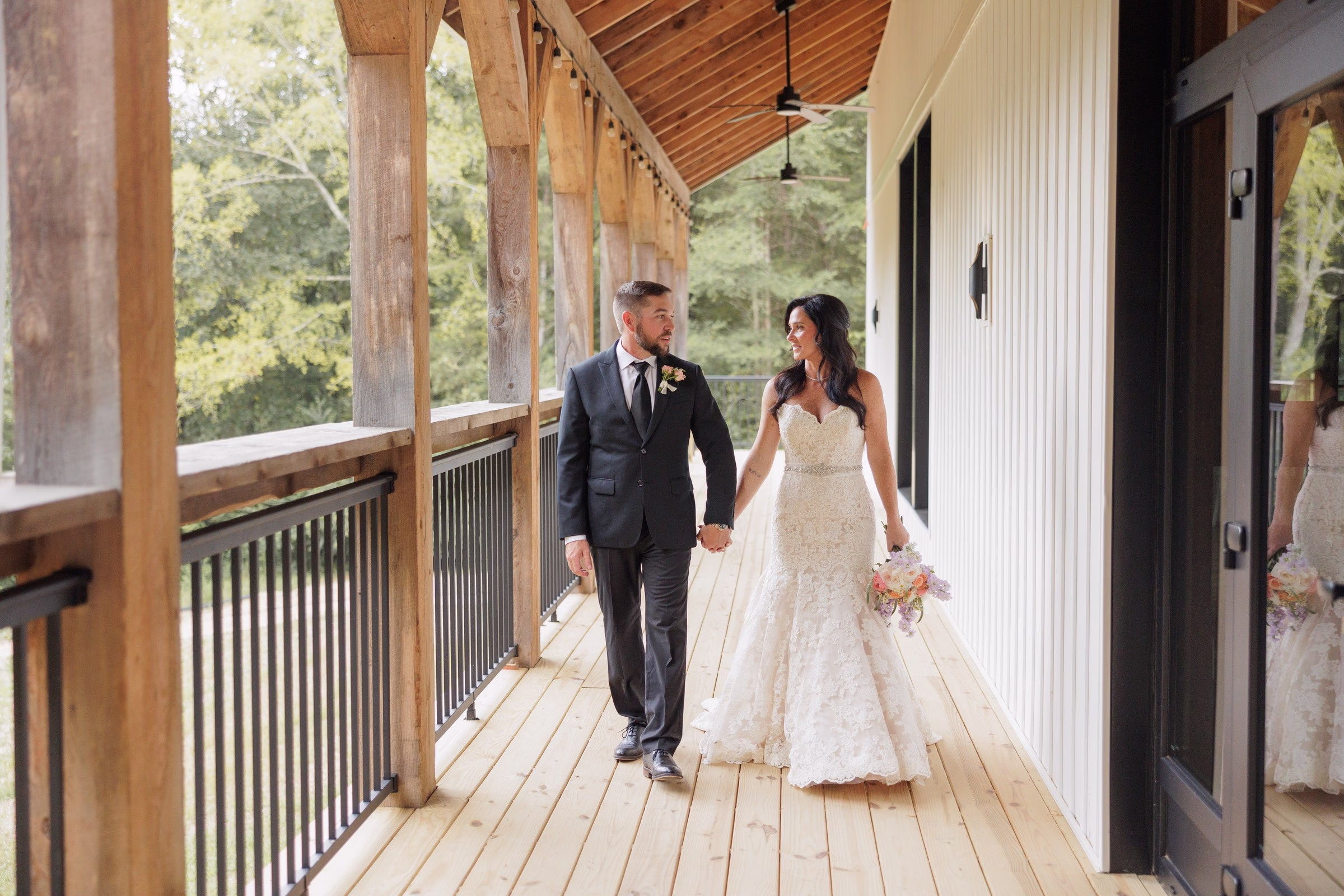Bride and groom walk hand in hand along the covered wooden porch at The Grand wedding venue in York SC near Charlotte NC, bride wearing a lace mermaid gown and holding a colorful bouquet