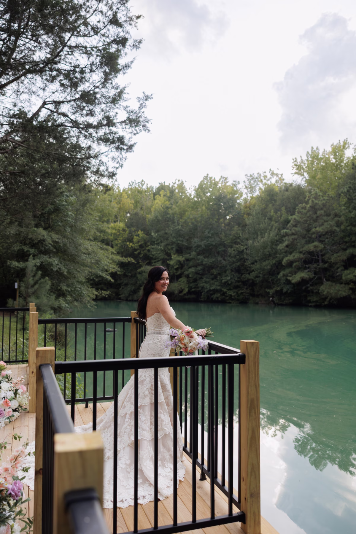 Bride in a strapless lace gown holding a pastel bouquet smiles over her shoulder while standing on a wooden deck overlooking the turquoise lake at The Grand wedding venue in York SC near Charlotte NC