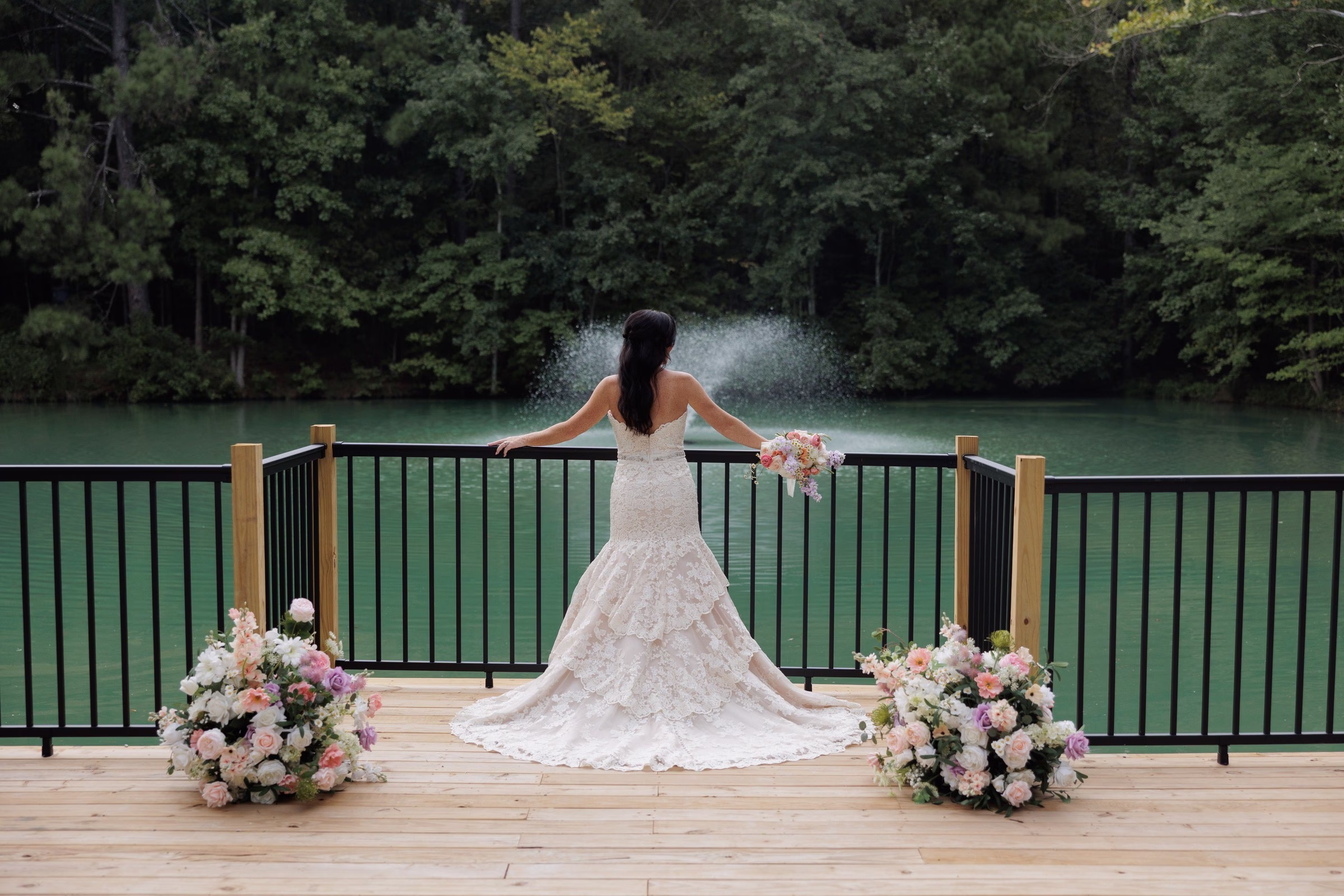 Bride in a lace mermaid gown holding a bridal bouquet stands on the lakeside deck overlooking a fountain at The Grand wedding venue in York SC near Charlotte NC