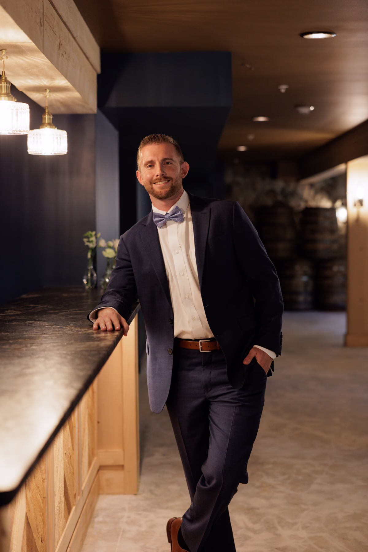Smiling groom in a navy suit and lavender bow tie leaning against the bar at The Grand wedding venue in York SC near Charlotte NC