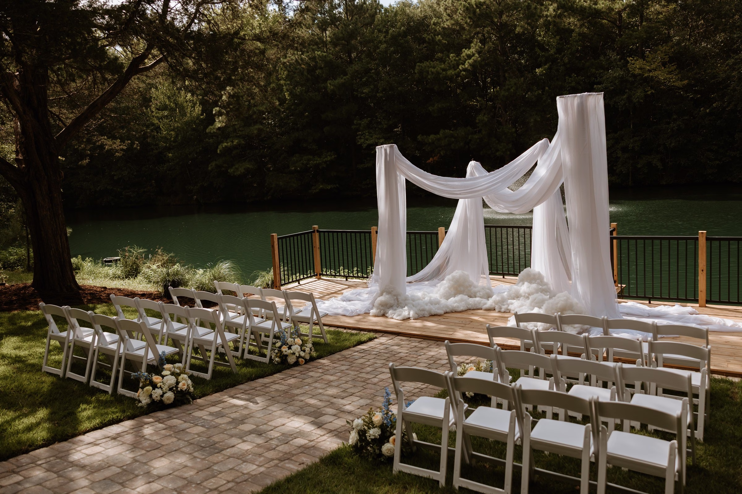 Elegant outdoor wedding ceremony setup at The Grand in York SC near Charlotte NC featuring a draped white fabric arch with cloud-like base decor on a wooden deck overlooking a green lake surrounded by trees with white folding chairs and floral arrangements lining the aisle