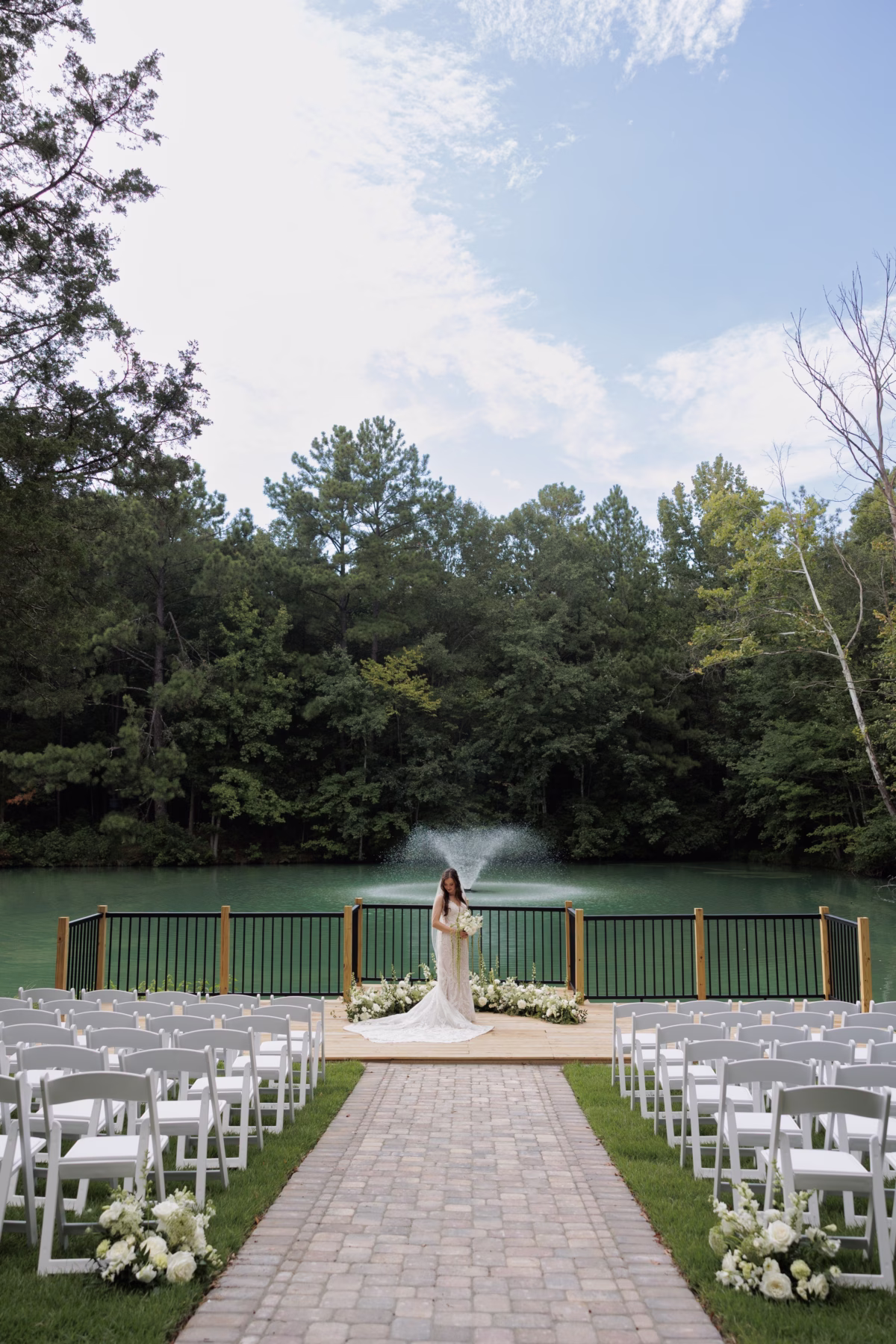 A bride stands at the altar of an elegantly arranged outdoor wedding ceremony with white chairs lining a brick aisle, white floral arrangements, and a fountain lake backdrop surrounded by lush trees at The Grand wedding venue in York SC near Charlotte NC