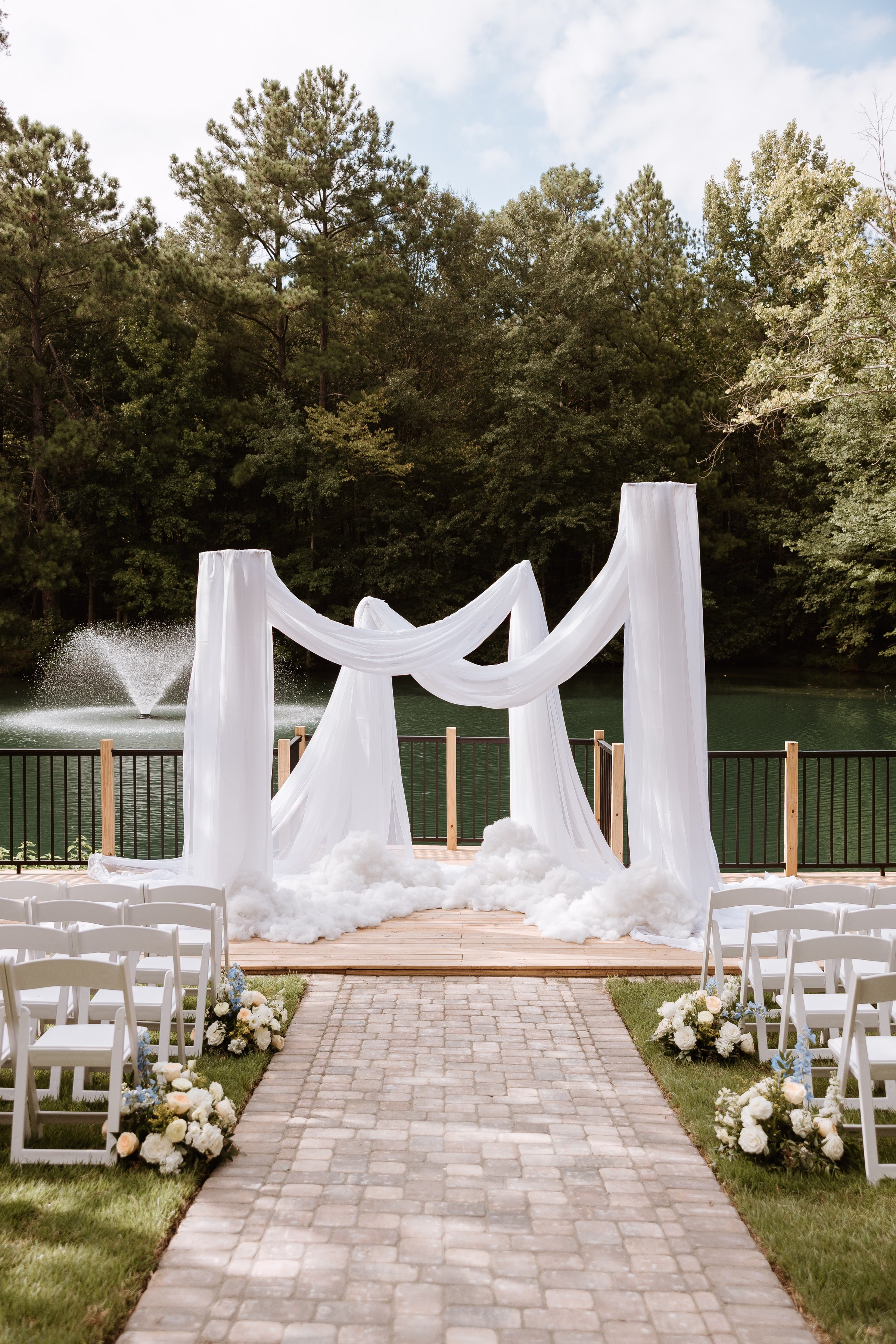 Elegant outdoor wedding ceremony setup at The Grand in York SC near Charlotte NC featuring a white draped arch on a wooden platform overlooking a lake with a fountain, flanked by white folding chairs and floral arrangements lining a brick aisle
