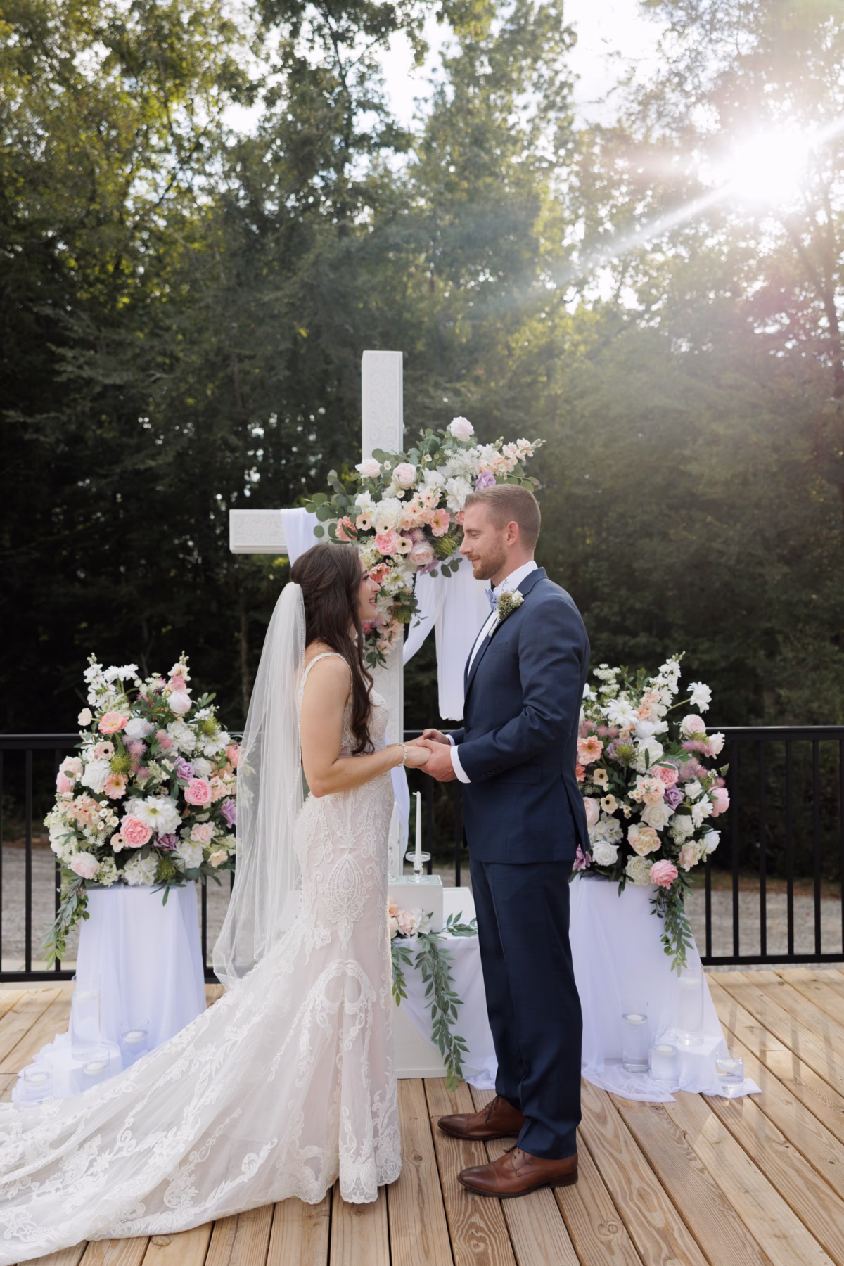 Bride and groom exchange vows beneath a floral-adorned white cross at The Grand wedding venue in York SC near Charlotte NC surrounded by lush pink and white floral arrangements