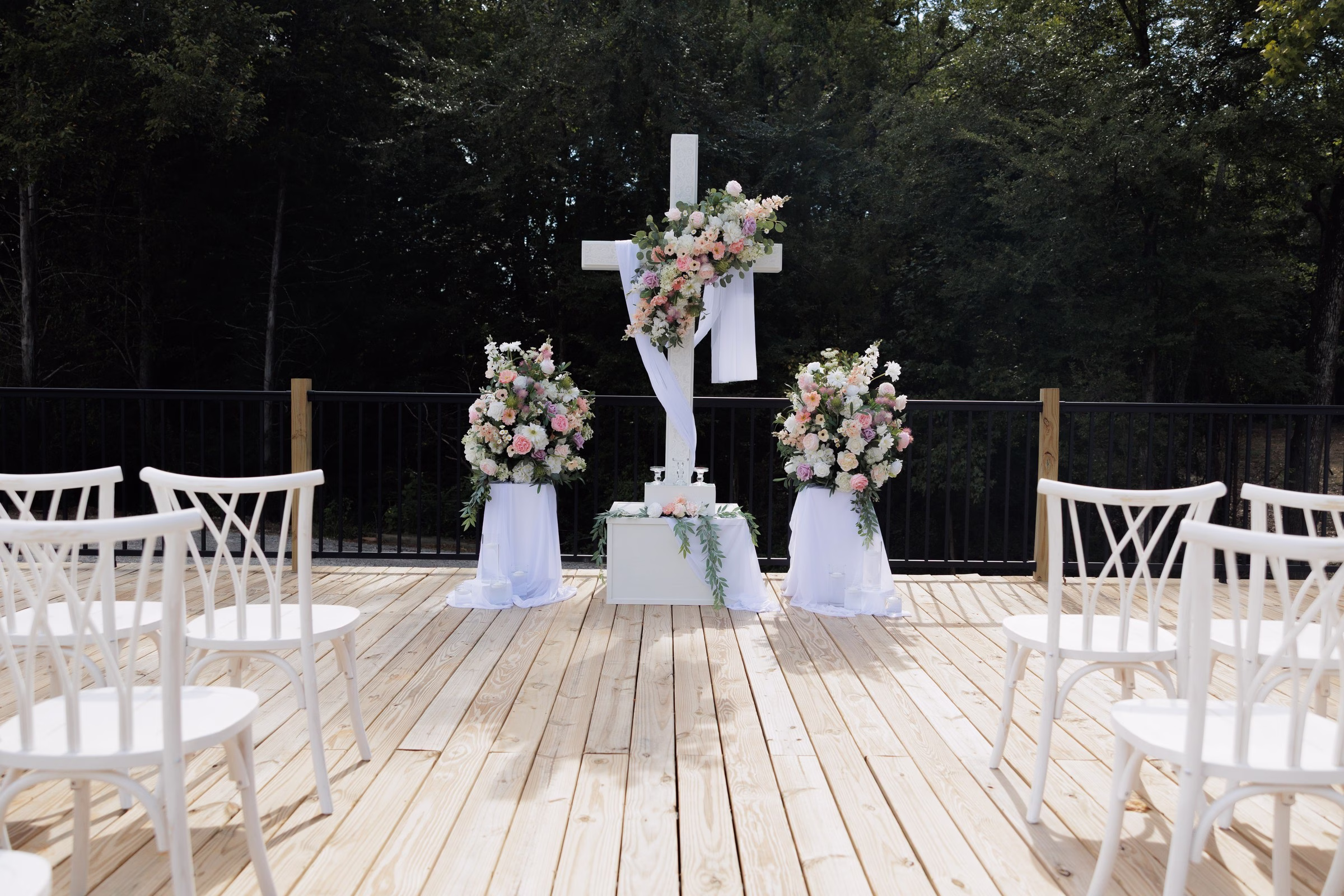 Elegant outdoor wedding ceremony setup at The Grand in York SC near Charlotte NC featuring a white cross adorned with pink and blush floral arrangements on a wooden deck surrounded by white chairs and lush forest backdrop