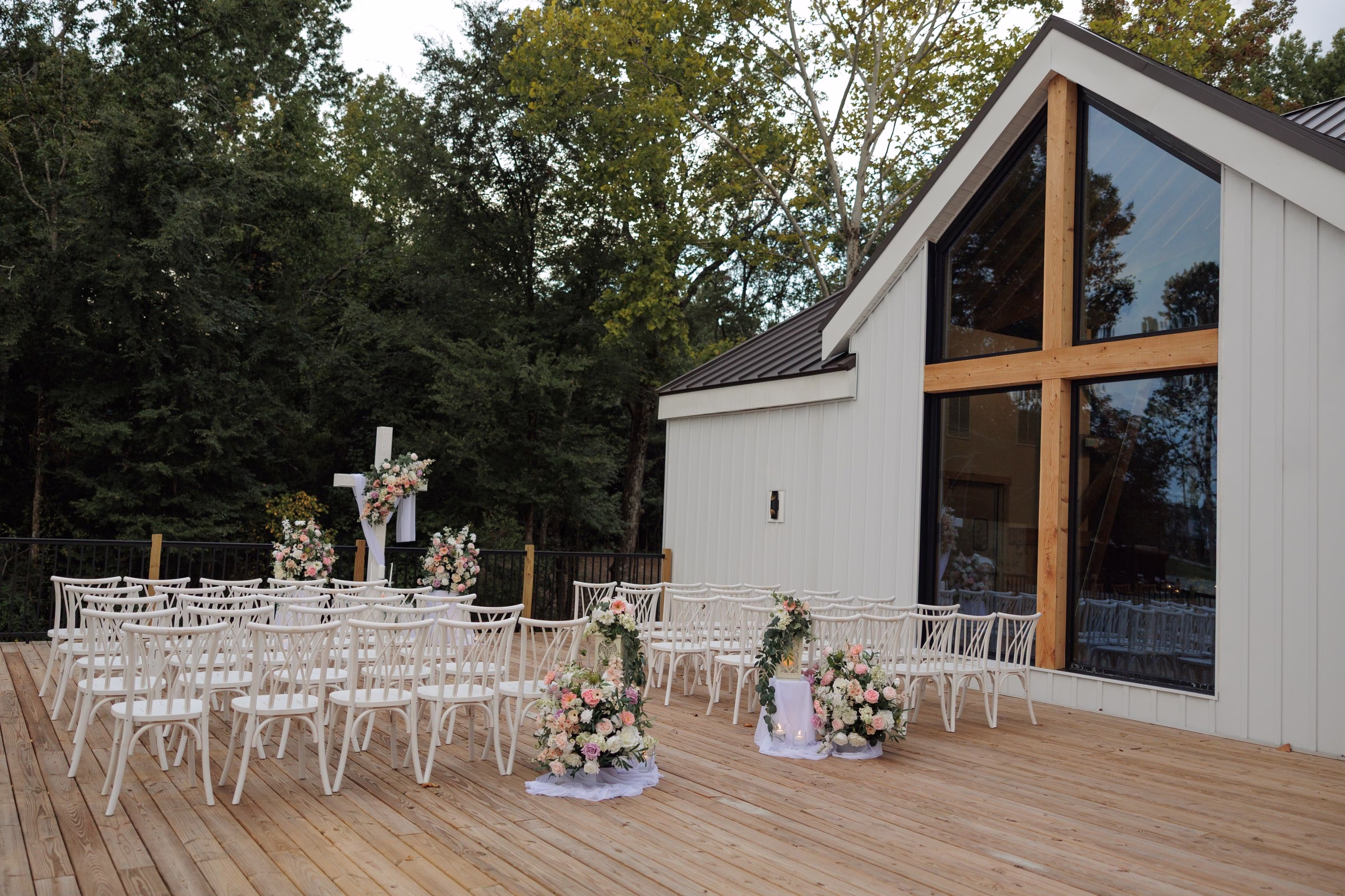 Outdoor wedding ceremony setup on a wooden deck at The Grand wedding venue in York SC near Charlotte NC featuring white chairs pink and white floral arrangements and a decorated cross altar backdrop