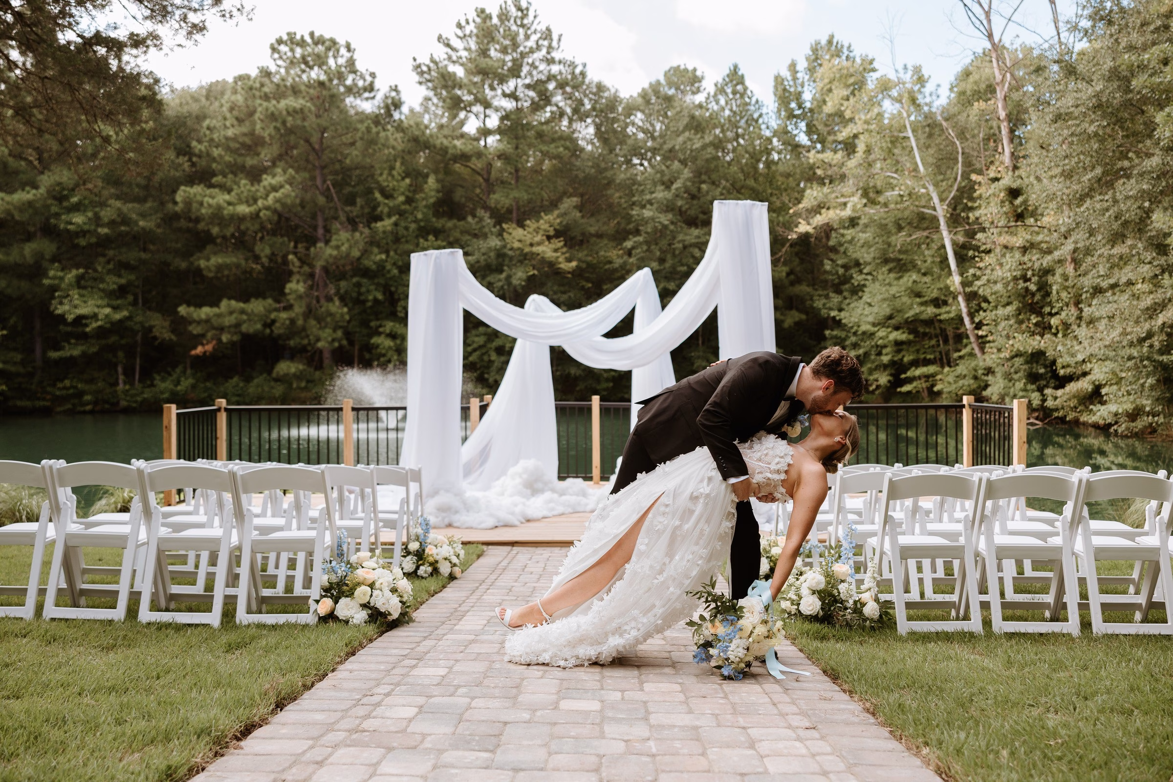 Bride and groom share a romantic dip kiss along the brick aisle at The Grand wedding venue in York SC near Charlotte NC with a white draped arch and pond backdrop