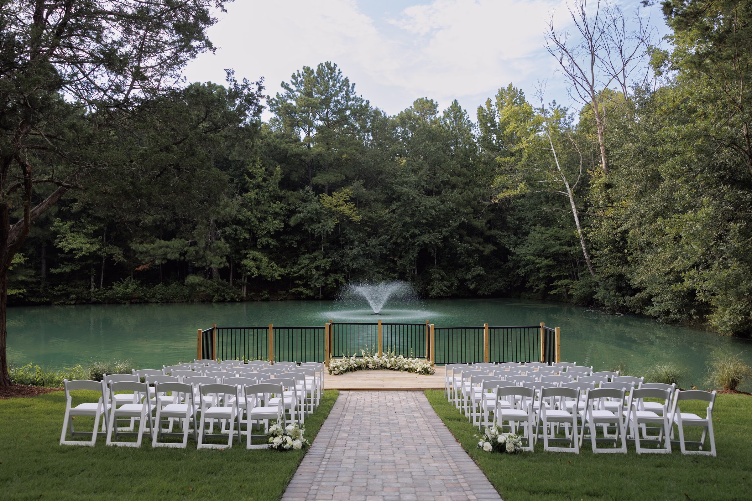 Outdoor wedding ceremony setup with white folding chairs lining a brick aisle leading to a floral altar on a deck overlooking a fountain lake at The Grand wedding venue in York SC near Charlotte NC