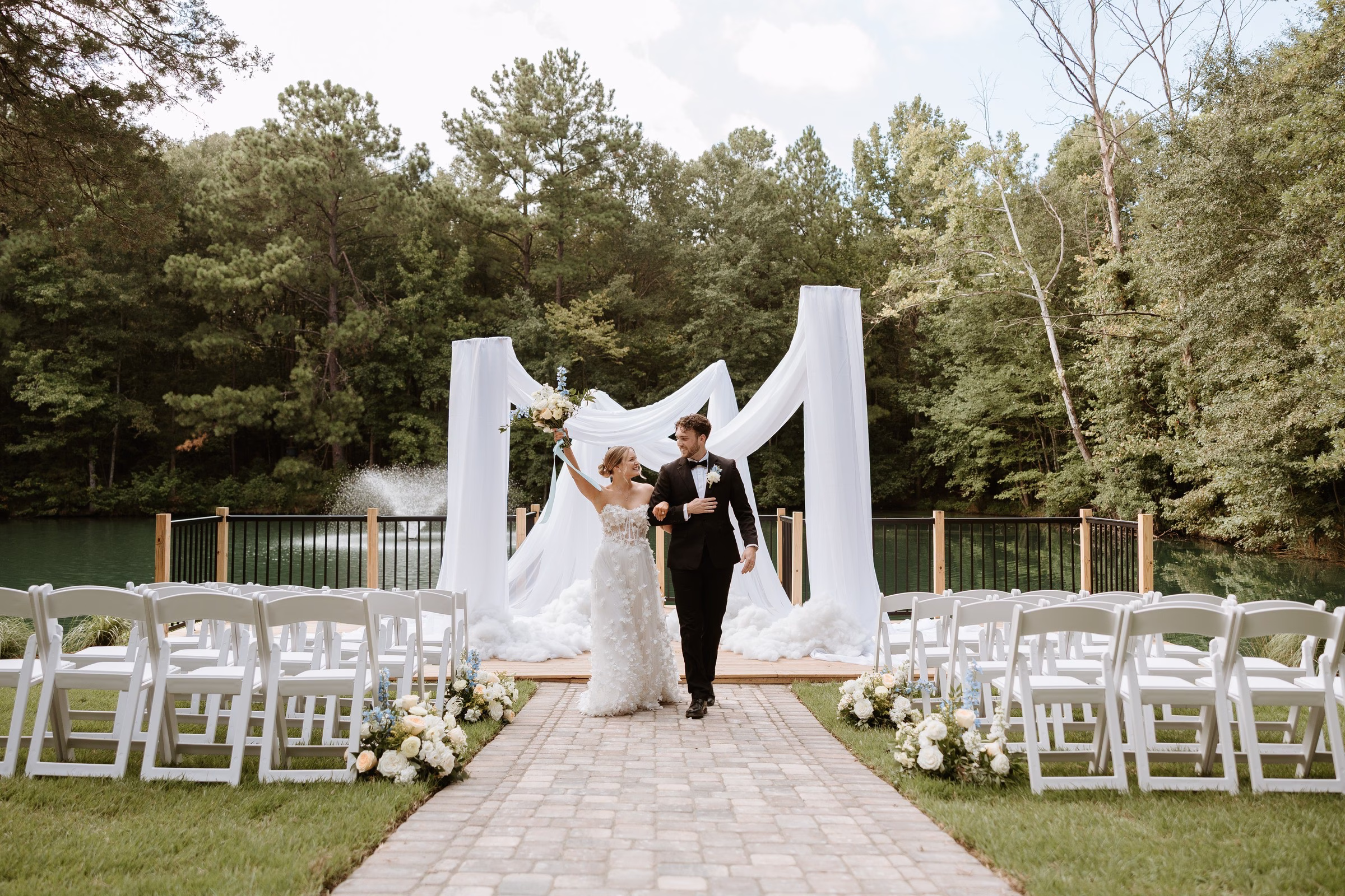 Bride and groom walking back down the aisle after their ceremony at The Grand wedding venue in York SC near Charlotte NC, surrounded by white draped arch, floral arrangements, and white folding chairs beside a scenic pond