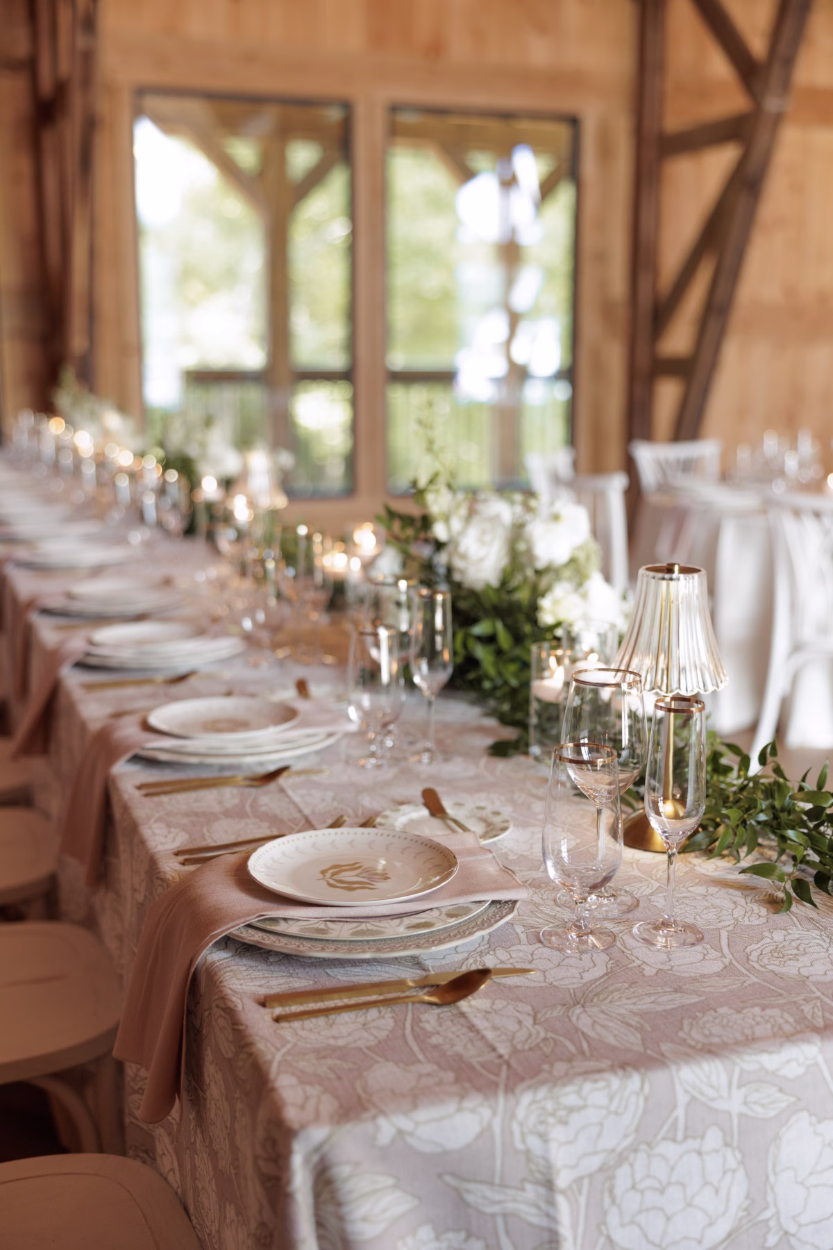 Elegant wedding reception table setting at The Grand in York SC near Charlotte NC featuring gold flatware floral patterned linens greenery garland white roses and gold accent candle lamps