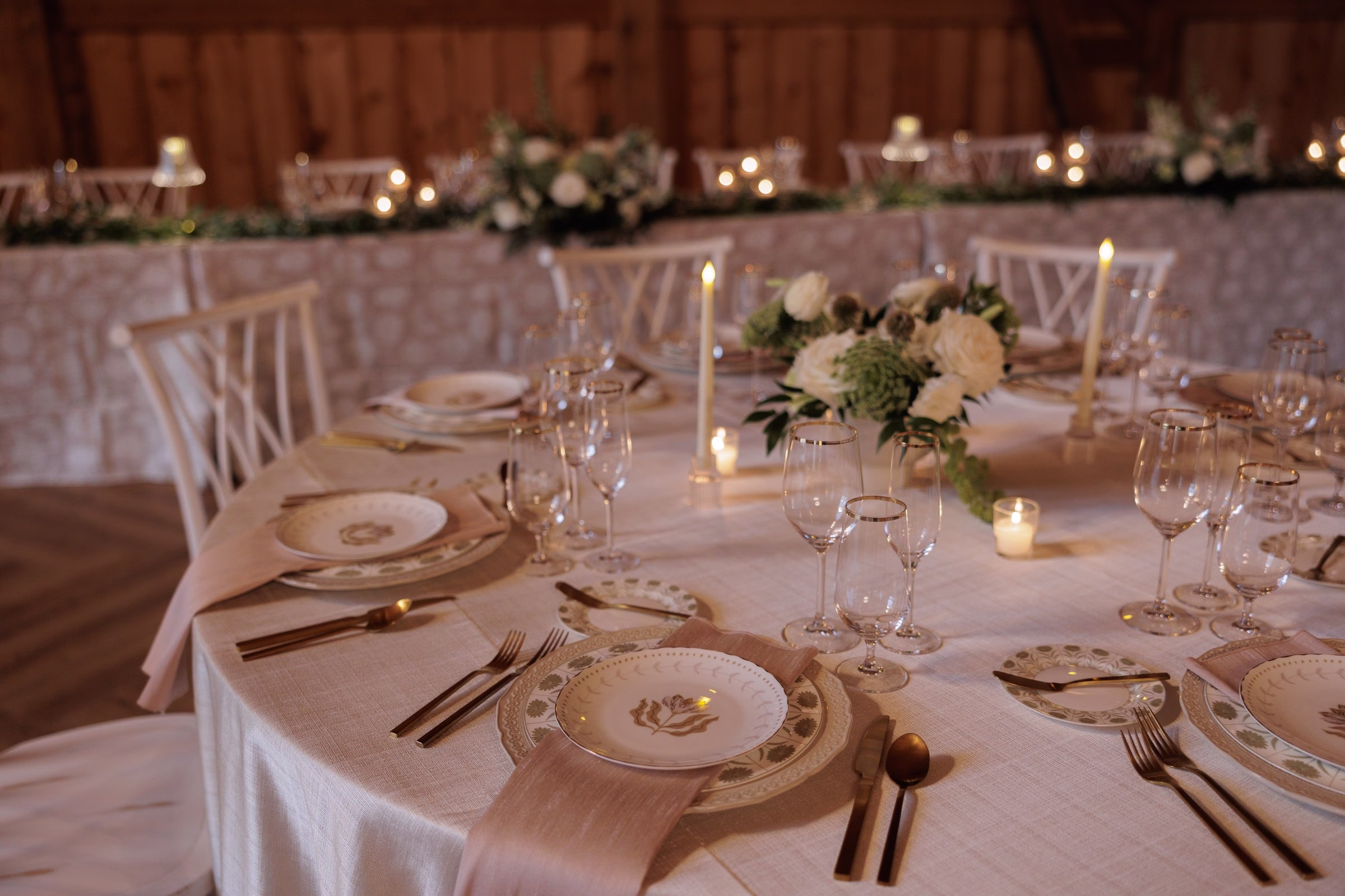 Elegant candlelit wedding reception table setting with gold-rimmed china, crystal stemware, white floral centerpiece, and blush linen napkins at The Grand wedding venue in York SC near Charlotte NC