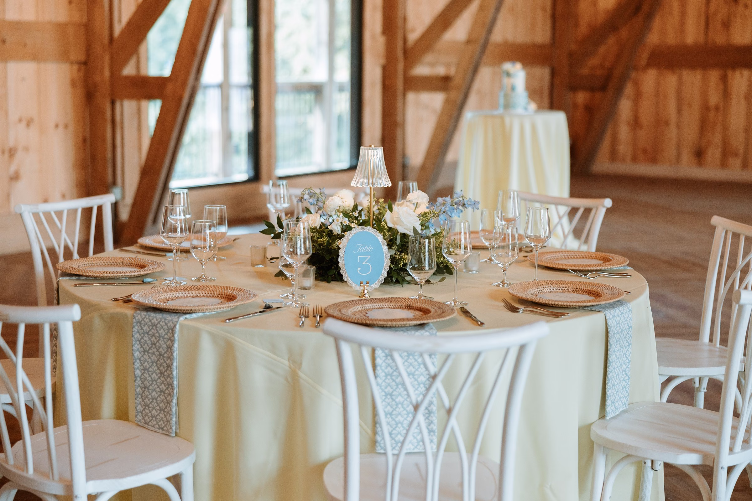 Elegantly styled wedding reception table number three with cream linens, blue patterned table runner, woven charger plates, crystal stemware, and a floral centerpiece of white roses and blue hydrangeas at The Grand wedding venue in York SC near Charlotte NC